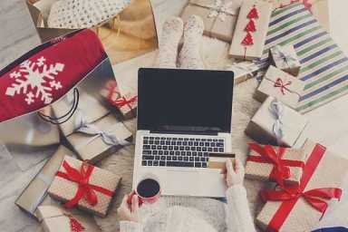 Person online shopping with laptop, coffee, and credit card, surrounded by holiday gifts and bags.