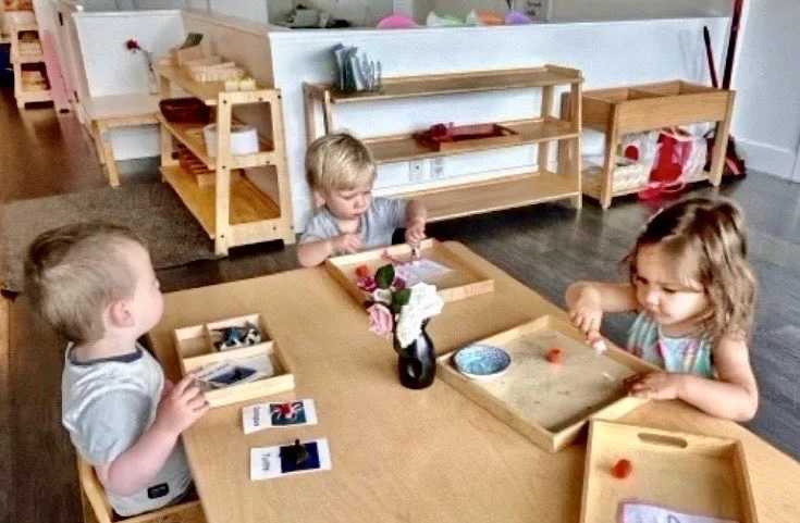 Three young children sit at a wooden table in a bright classroom, engaged in hands-on activities with trays, cards, and small objects.