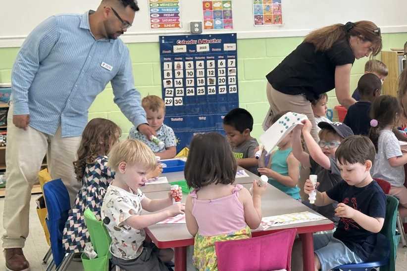 A group of preschool children sit at colorful tables, using glue sticks and crafts, supervised by two adults in a bright classroom setting.