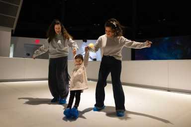 Two women hold hands with a young child as they all glide around the New York Hall of Science new experience, Space Glide