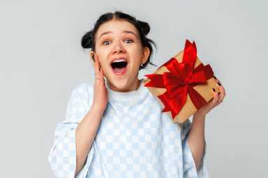 Portrait of cheerful amazed young dark-haired girl holding in hand gift having fun, standing in trendy t shirt over light grey background. Teenage girls in love, Receiving gifts from lovers