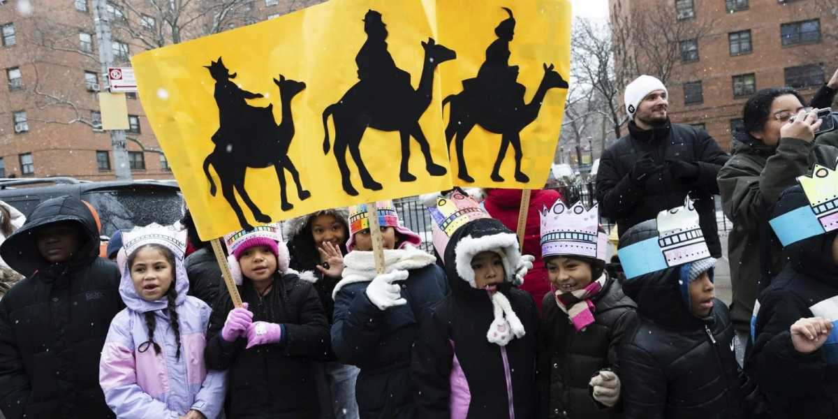 Children lined up wearing winter coats and hats and holding up yellow posters with silhouettes of the Three Wise Men on camels to celebrate Three Kings Day.