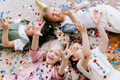 4 laughing children lying on a rug reaching their hands up as confetti falls on them