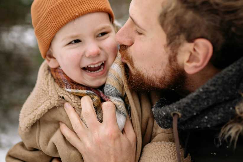 Little boy wearing a winter hat and coat being kissed on the cheek by his father