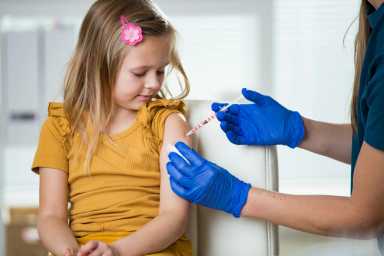 Female nurse with surgical mask and in gloves giving childhood vaccine injection to a young girl in clinic. Children vaccination.