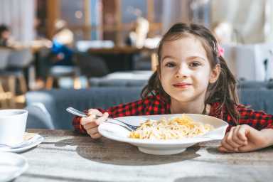 Smiling girl 6-7 years old sitting at a table with a large plate of spaghetti with cheese at restaurant week