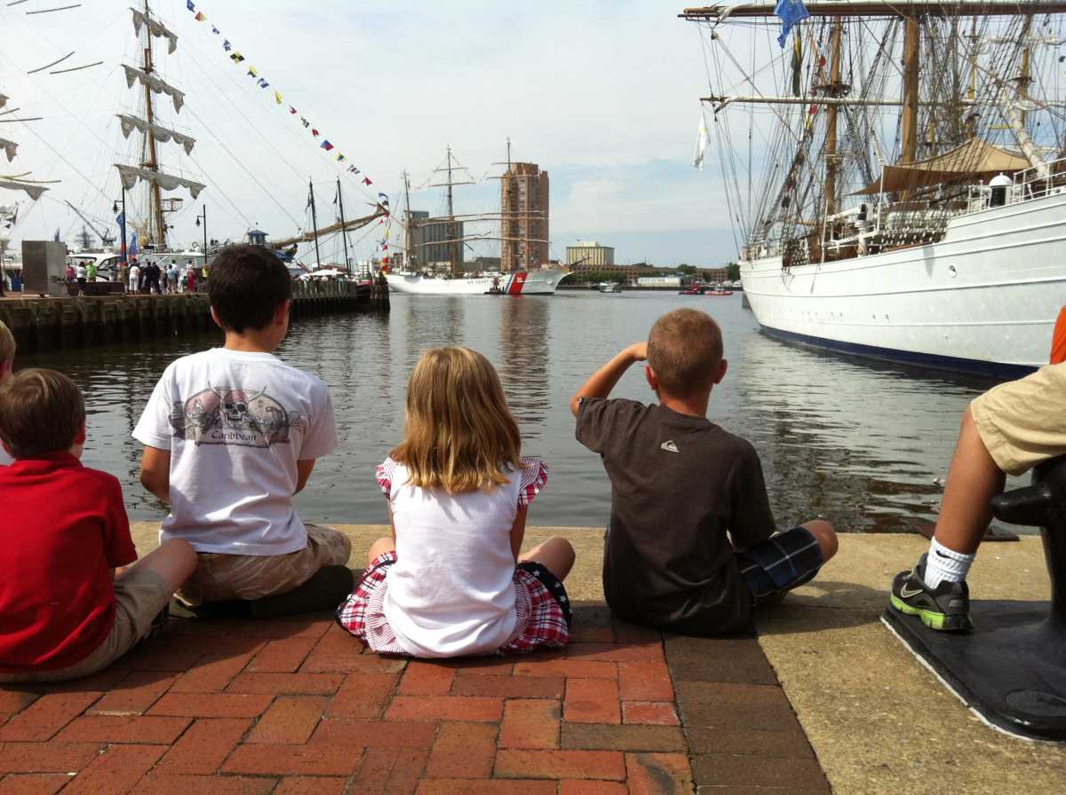 Children sitting on a dock with their backs turned to the camera, looking ahead at the ships on either side of them, Sail4th