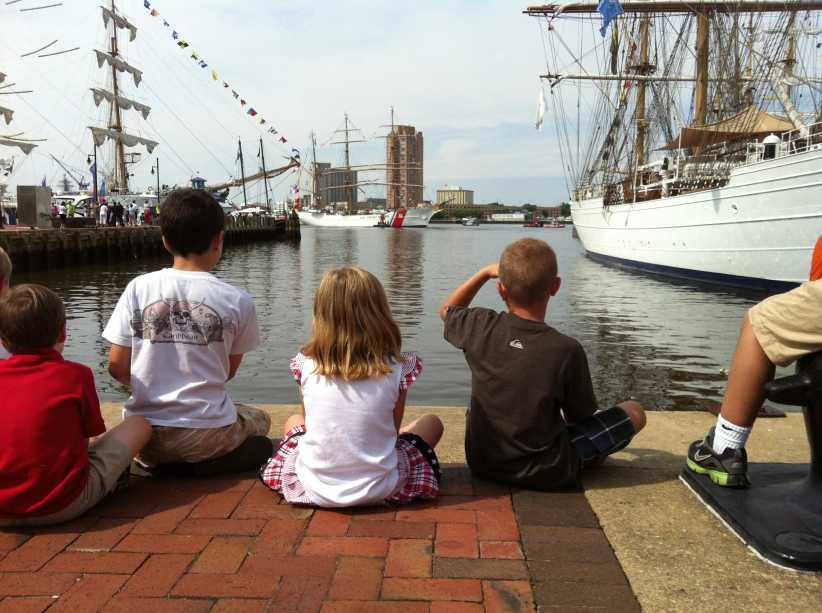 Children sitting on a dock with their backs turned to the camera, looking ahead at the ships on either side of them, Sail4th