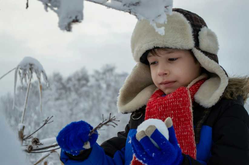 Little boy wearing. a big winter hat and holding a snowball