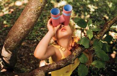 Young blonde girl looking up at a tree with homemade binoculars.