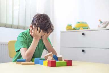 a 7-9-year-old boy in a green T-shirt plays with wooden blocks in the children's room having a meltdown. He covers his face with his hands. Refusing to play. Protest.