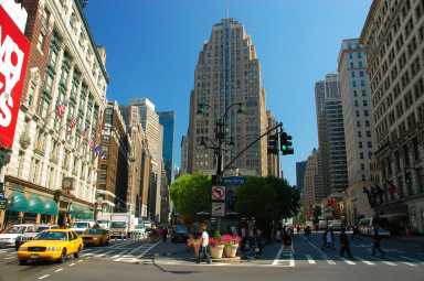 A new TJ Maxx store is opening at Herald Square, at 34th street looking at Broadway street (left) and Avenue of the Americas (right), with many pedestrians walking (and some standing), as well as traffic on Broadway (including taxi cabs.)