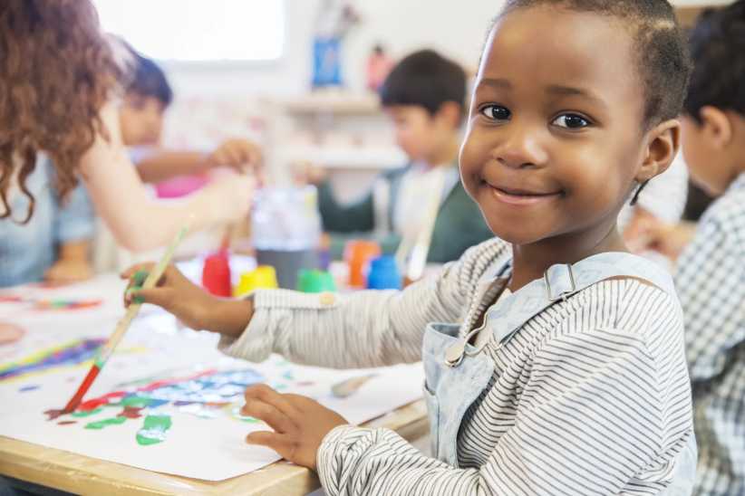 Pre K student painting in class, looking at the camera and smiling